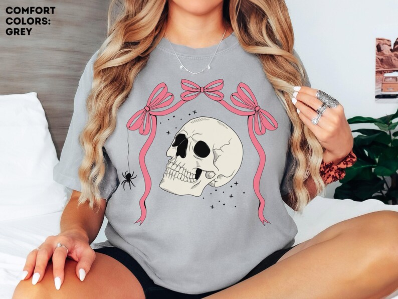 K&ouml;nnte beinhalten: Graues T-Shirt mit einem Totenkopf-Motiv. Der Totenkopf ist cremefarben mit schwarzen Augenh&ouml;hlen und einem detaillierten Kiefer. Rosa Schleifen und B&auml;nder umrahmen den Totenkopf, mit einer Spinne, die an einem Band h&auml;ngt. Der Text "COMFORT COLORS: GREY" steht oben links.