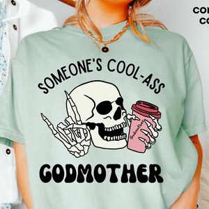 May include: A light green t-shirt featuring a graphic of a skeleton holding a coffee cup with the text "Godmother" and the phrase "Someone's Cool-Ass Godmother" printed on it.
