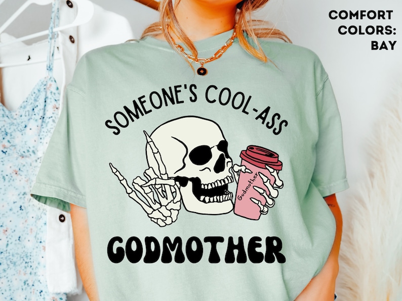 May include: A light green t-shirt featuring a graphic of a skeleton holding a coffee cup with the text "Godmother" and the phrase "Someone's Cool-Ass Godmother" printed on it.