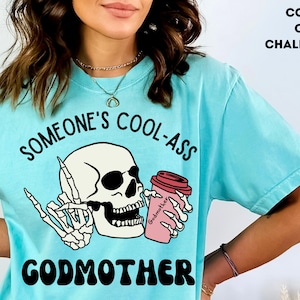 May include: A light blue t-shirt featuring a skull wearing sunglasses and holding a pink coffee cup with the text "Godmother" on it. The text "Someone's Cool-Ass Godmother" is printed on the shirt.