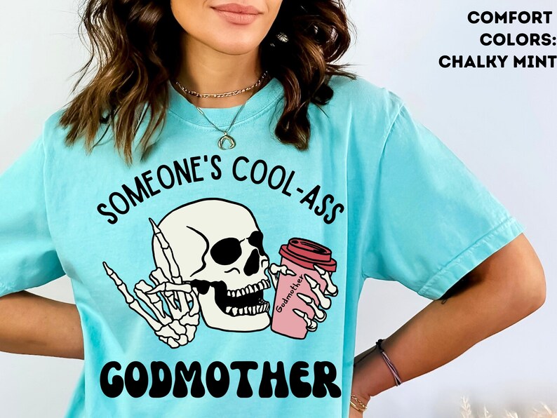 May include: A light blue t-shirt featuring a skull wearing sunglasses and holding a pink coffee cup with the text "Godmother" on it. The text "Someone's Cool-Ass Godmother" is printed on the shirt.
