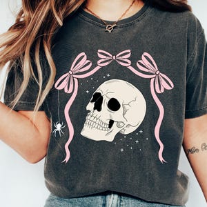K&ouml;nnte beinhalten: Dunkelgraues T-Shirt mit einem cremefarbenen Totenkopf-Design, schwarzen Augenh&ouml;hlen und Z&auml;hnen, rosa Schleifen und B&auml;ndern. Eine wei&szlig;e Spinne h&auml;ngt von dem Design. Das Shirt hat den Text "COMFORT COLORS: PEPPER".