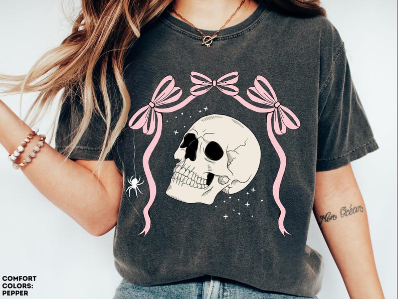 K&ouml;nnte beinhalten: Dunkelgraues T-Shirt mit einem cremefarbenen Totenkopf-Design, schwarzen Augenh&ouml;hlen und Z&auml;hnen, rosa Schleifen und B&auml;ndern. Eine wei&szlig;e Spinne h&auml;ngt von dem Design. Das Shirt hat den Text "COMFORT COLORS: PEPPER".