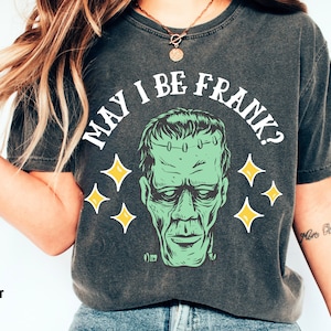 May include: Dark gray t-shirt with the phrase "MAY I BE FRANK?" above a green Frankenstein's monster graphic. The design includes yellow star accents. The shirt is a comfort color in pepper.