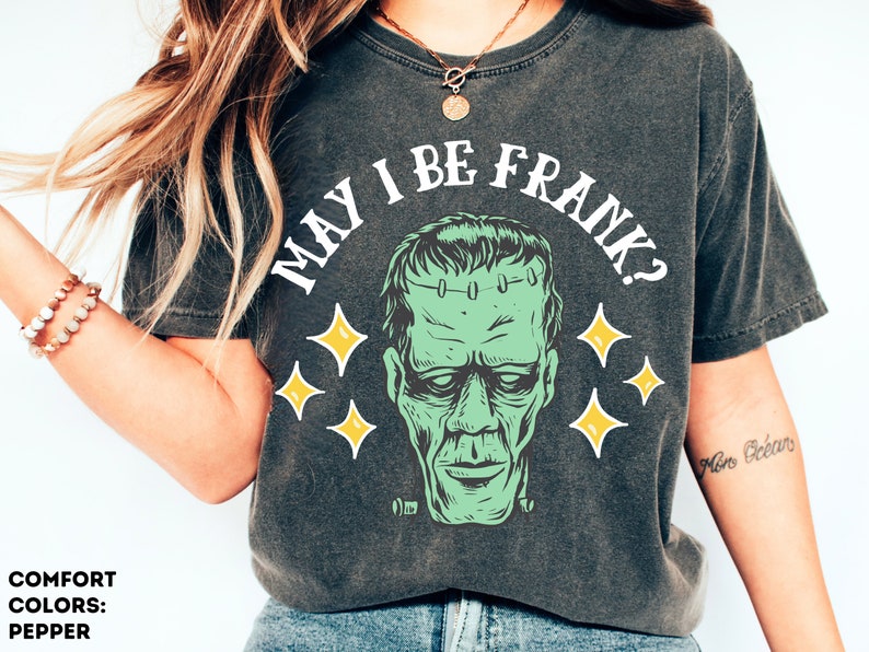 May include: Dark gray t-shirt with the phrase "MAY I BE FRANK?" above a green Frankenstein's monster graphic. The design includes yellow star accents. The shirt is a comfort color in pepper.