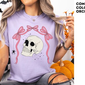 K&ouml;nnte beinhalten: Lilafarbenes T-Shirt mit einem Totenkopf-Design, rosa Schleifen und einer Spinne. Der Text "COMFORT COLORS: ORCHID" befindet sich oben rechts. Dieses Halloween-T-Shirt ist Teil einer Kollektion gruseliger Kleidung.