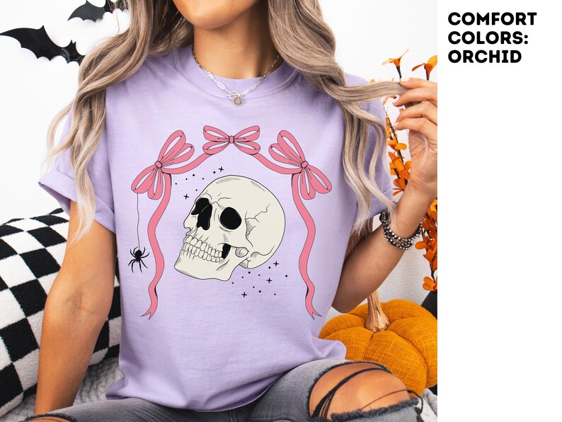 K&ouml;nnte beinhalten: Lilafarbenes T-Shirt mit einem Totenkopf-Design, rosa Schleifen und einer Spinne. Der Text "COMFORT COLORS: ORCHID" befindet sich oben rechts. Dieses Halloween-T-Shirt ist Teil einer Kollektion gruseliger Kleidung.