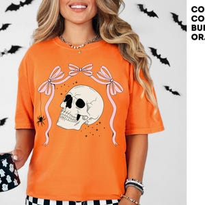 K&ouml;nnte beinhalten: Burnt-Orange-T-Shirt mit Totenkopf-Grafik, rosa Schleifen und einer Spinne. Auf dem Shirt steht der Text "COMFORT COLORS: BURNT ORANGE". Eine schwarz-wei&szlig;e Tasse mit Geistermotiv wird gehalten. Schwarze Fledermausdekorationen sind im Hintergrund.