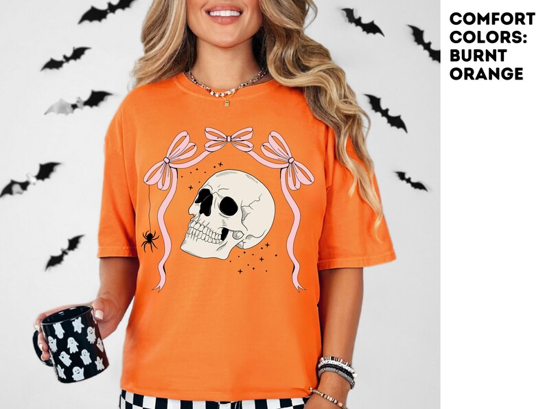 K&ouml;nnte beinhalten: Burnt-Orange-T-Shirt mit Totenkopf-Grafik, rosa Schleifen und einer Spinne. Auf dem Shirt steht der Text "COMFORT COLORS: BURNT ORANGE". Eine schwarz-wei&szlig;e Tasse mit Geistermotiv wird gehalten. Schwarze Fledermausdekorationen sind im Hintergrund.