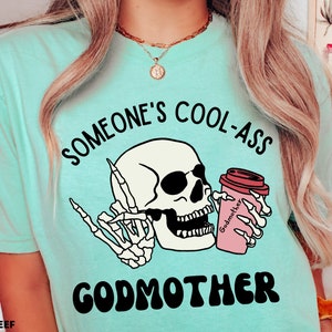May include: A turquoise blue t-shirt with the text "Someone's Cool-Ass Godmother" in black and white. The design features a cartoon skull with sunglasses holding a pink coffee cup with the word "Godmother" on it.