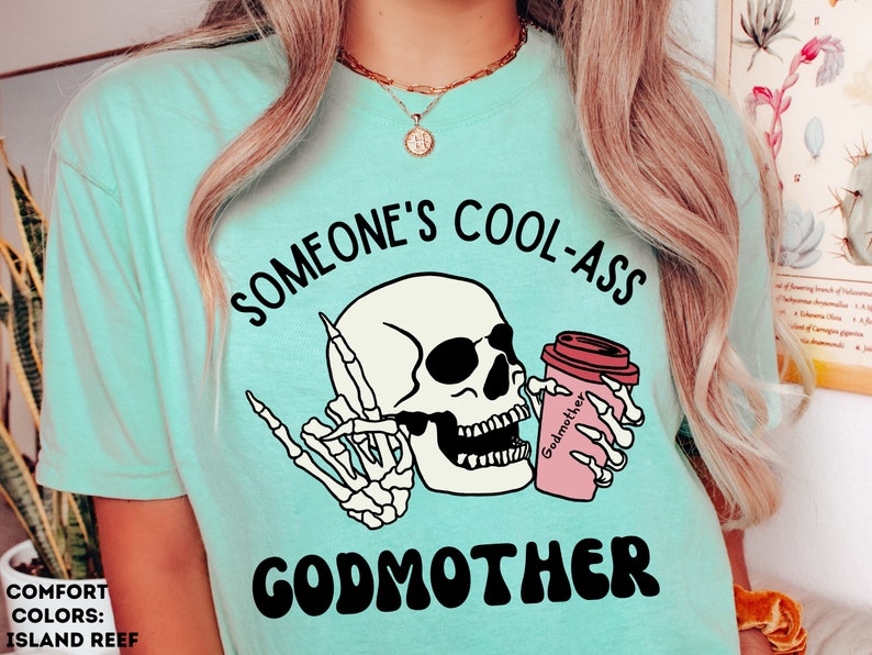 May include: A turquoise blue t-shirt with the text "Someone's Cool-Ass Godmother" in black and white. The design features a cartoon skull with sunglasses holding a pink coffee cup with the word "Godmother" on it.