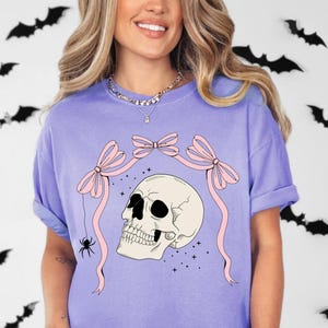 K&ouml;nnte beinhalten: Violettes Comfort Colors T-Shirt mit einem Totenkopf-Design, rosa Schleifen und einer Spinne. Der Totenkopf ist cremefarben mit schwarzen Augenh&ouml;hlen. Schwarze Fledermaus-Ausschnitte schm&uuml;cken den Hintergrund. Das Shirt hat eine lockere Passform.