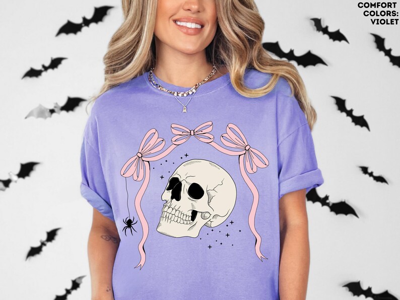 K&ouml;nnte beinhalten: Violettes Comfort Colors T-Shirt mit einem Totenkopf-Design, rosa Schleifen und einer Spinne. Der Totenkopf ist cremefarben mit schwarzen Augenh&ouml;hlen. Schwarze Fledermaus-Ausschnitte schm&uuml;cken den Hintergrund. Das Shirt hat eine lockere Passform.