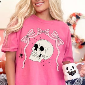 K&ouml;nnte beinhalten: Rosafarbenes T-Shirt mit Totenkopf-Grafik, Schleifen und einer Spinne. Der Text "COMFORT COLORS: CRUNCH BERRY" befindet sich oben links. Die Person h&auml;lt einen wei&szlig;en Becher mit einem Geistergesicht.