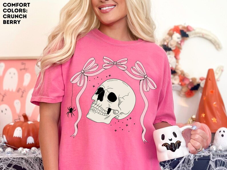 K&ouml;nnte beinhalten: Rosafarbenes T-Shirt mit Totenkopf-Grafik, Schleifen und einer Spinne. Der Text "COMFORT COLORS: CRUNCH BERRY" befindet sich oben links. Die Person h&auml;lt einen wei&szlig;en Becher mit einem Geistergesicht.