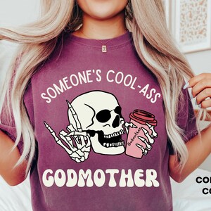 May include: Purple t-shirt featuring a white skull wearing sunglasses and holding a pink coffee cup with the text "Godmother" and the phrase "Someone's Cool-Ass Godmother". The text "Comfort Colors: Berry" is visible in the bottom right corner.