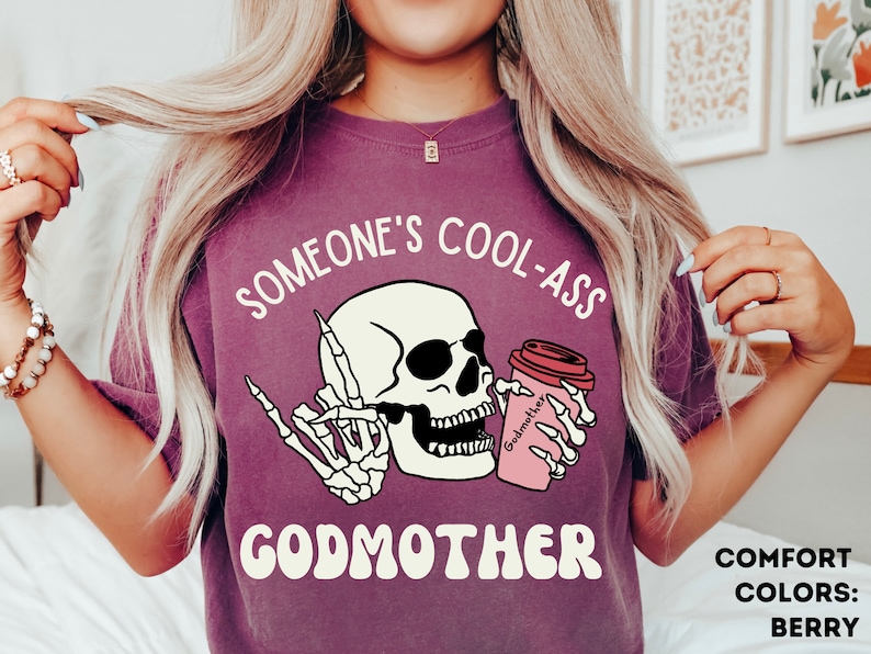 May include: Purple t-shirt featuring a white skull wearing sunglasses and holding a pink coffee cup with the text "Godmother" and the phrase "Someone's Cool-Ass Godmother". The text "Comfort Colors: Berry" is visible in the bottom right corner.