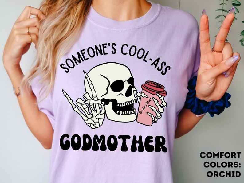 May include: A purple t-shirt with the text "Someone's Cool-Ass Godmother" in black lettering. A cartoon skull with sunglasses is holding a pink coffee cup with the text "Godmother" on it.