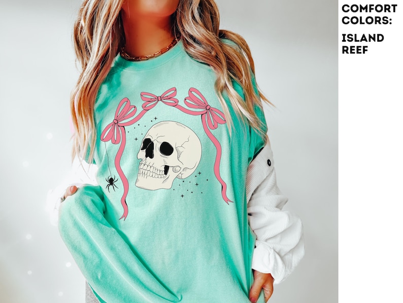 K&ouml;nnte beinhalten: Ein hellt&uuml;rkisfarbenes Langarmshirt mit einem Totenkopf-Design, rosa Schleifen und einer Spinne. Das Shirt hat den Text "COMFORT COLORS: ISLAND REEF" in der oberen rechten Ecke.