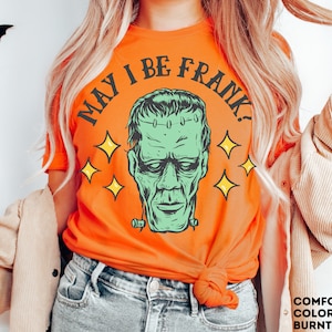 May include: Burnt orange t-shirt featuring a green Frankenstein's monster graphic with the text "MAY I BE FRANK?" and yellow star accents. The shirt is a comfort color and is perfect for Halloween.
