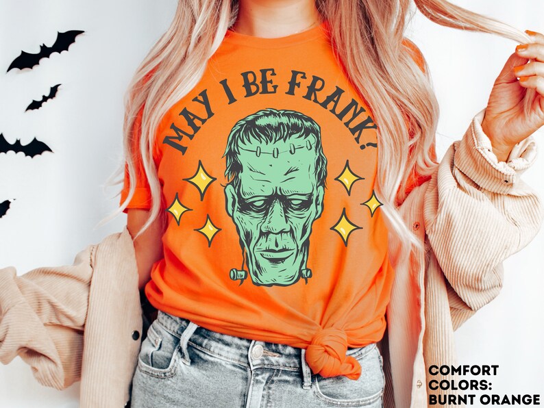 May include: Burnt orange t-shirt featuring a green Frankenstein's monster graphic with the text "MAY I BE FRANK?" and yellow star accents. The shirt is a comfort color and is perfect for Halloween.