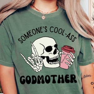 May include: Green t-shirt featuring a skull wearing sunglasses and holding a coffee cup that reads "Godmother". The text on the shirt says "Someone's Cool-Ass Godmother".