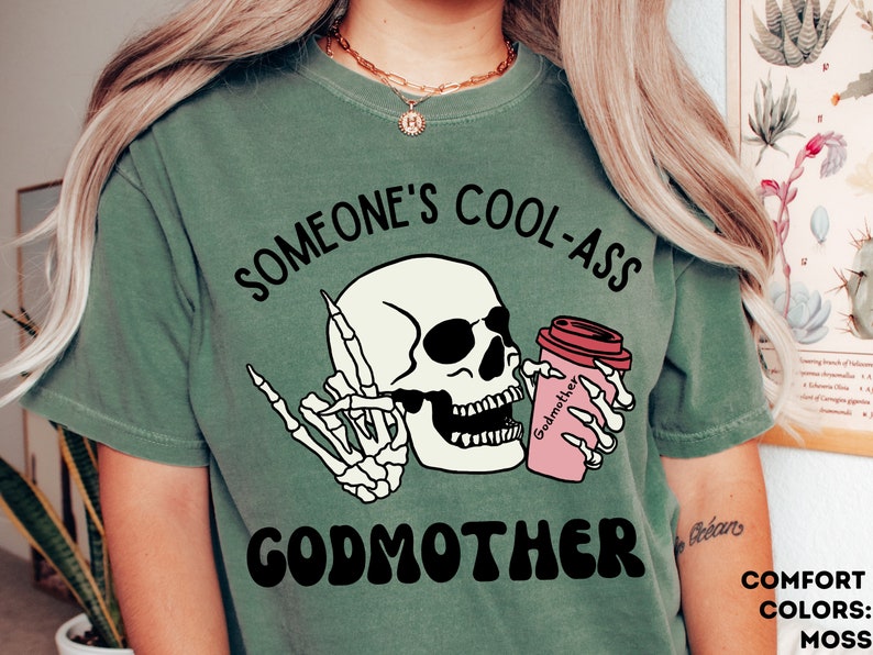 May include: Green t-shirt featuring a skull wearing sunglasses and holding a coffee cup that reads "Godmother". The text on the shirt says "Someone's Cool-Ass Godmother".