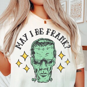May include: Ivory-colored t-shirt featuring a green Frankenstein's monster graphic with the text "MAY I BE FRANK?" in black. The design includes yellow star accents, creating a Halloween or spooky theme.