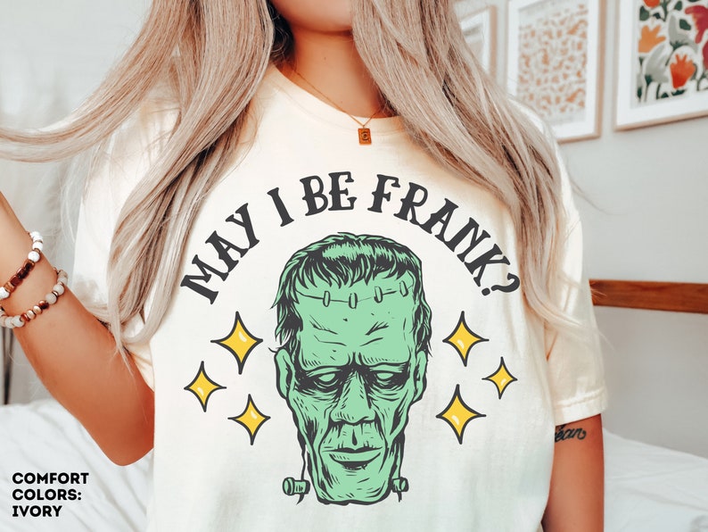 May include: Ivory-colored t-shirt featuring a green Frankenstein's monster graphic with the text "MAY I BE FRANK?" in black. The design includes yellow star accents, creating a Halloween or spooky theme.