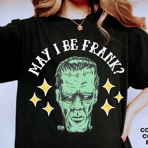 May include: Black t-shirt featuring a green Frankenstein's monster graphic with the text "MAY I BE FRANK?" in white. The design includes yellow starbursts. The shirt's color is black.