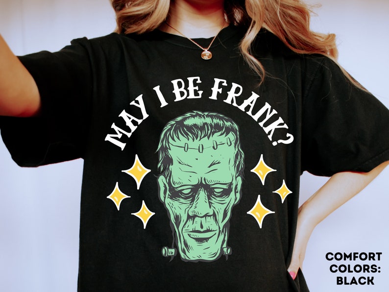 May include: Black t-shirt featuring a green Frankenstein's monster graphic with the text "MAY I BE FRANK?" in white. The design includes yellow starbursts. The shirt's color is black.