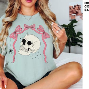 K&ouml;nnte beinhalten: Hellgr&uuml;nes T-Shirt mit einem Totenkopf-Design, rosa Schleifen und einer Spinne. Der Sch&auml;del hat schwarze Augenh&ouml;hlen und ist von Sternen umgeben. Der Text "COMFORT COLORS: BAY" befindet sich in der oberen rechten Ecke.