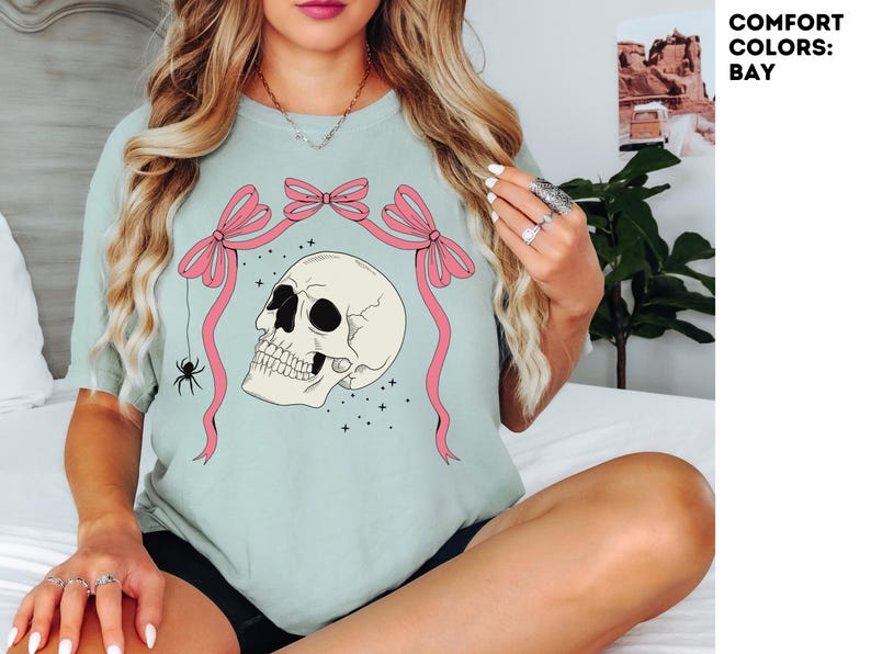 K&ouml;nnte beinhalten: Hellgr&uuml;nes T-Shirt mit einem Totenkopf-Design, rosa Schleifen und einer Spinne. Der Sch&auml;del hat schwarze Augenh&ouml;hlen und ist von Sternen umgeben. Der Text "COMFORT COLORS: BAY" befindet sich in der oberen rechten Ecke.
