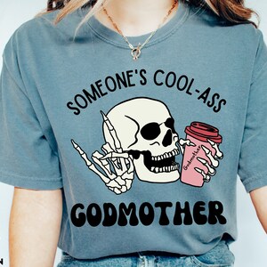 May include: A blue t-shirt with the text "Someone's Cool-Ass Godmother" in black and white. A cartoon skeleton holding a pink coffee cup with the word "Godmother" on it is in the centre of the shirt.