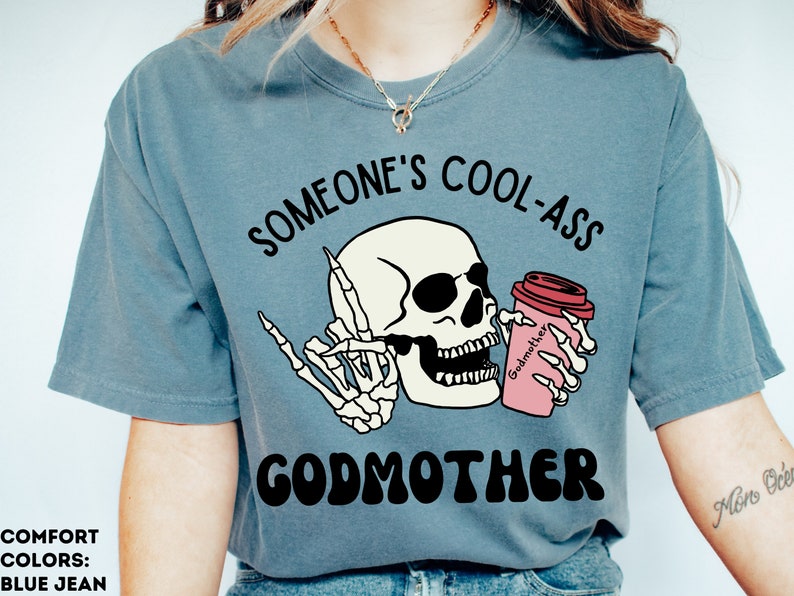 May include: A blue t-shirt with the text "Someone's Cool-Ass Godmother" in black and white. A cartoon skeleton holding a pink coffee cup with the word "Godmother" on it is in the centre of the shirt.