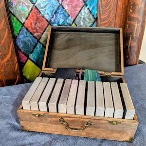 May include: A rectangular wooden storage box with a hinged lid and a metal handle, containing a row of ten upright, silver-toned rectangular cases. Behind the cases, several thin, colourful, translucent dividers are visible inside the box.