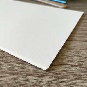 May include: A stack of rectangular, off-white paper sheets with a textured surface. The paper is positioned at an angle, showing the layered edges of the individual sheets. The surface of the paper appears matte and slightly fibrous in texture.