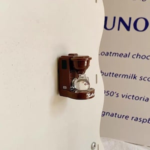 May include: A miniature brown coffee maker with a glass carafe. The small appliance is mounted on a white surface. The word "Cubs" is visible on the base of the coffee maker.