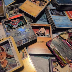 Könnte beinhalten: Eine Sammlung von geschichteten 3D-Schattenbox-Sammelkarten mit verschiedenen Charakteren wie Hawlucha, Pancham und Eevee. Jede Karte zeigt detaillierte Grafiken, Charakternamen und Spielstatistiken, wobei mehrere Stapel in einem Layout angeordnet sind.
