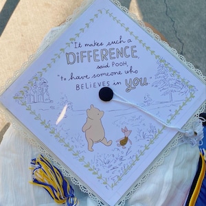 May include: A graduation cap with a Winnie the Pooh theme. The cap features a quote: "It makes such a DIFFERENCE, said POOH, to have someone who BELIEVES in you." The design includes illustrations of Winnie the Pooh and Piglet, with a tassel.