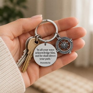May include: A silver-toned metal keychain featuring a round pendant engraved with the text, "In all your ways acknowledge him, and he shall direct your path. Proverbs 3:6," alongside a compass charm and two standard brass-colored keys.