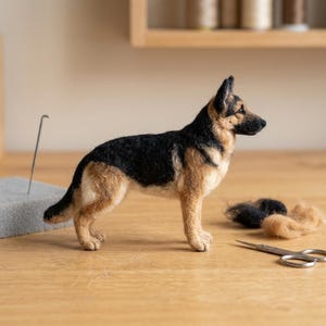 May include: A needle-felted German Shepherd dog figurine featuring a black and tan coat. The sculpture stands in a profile position, showcasing detailed texture and realistic colouring. Nearby are small tufts of loose wool and metal scissors.