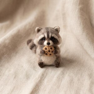 May include: A needle-felted raccoon figurine crafted from grey, white, and black wool fibers. The small creature features a striped tail, dark eyes, and a black nose, while holding a round, tan-colored cookie with dark brown chocolate chips.