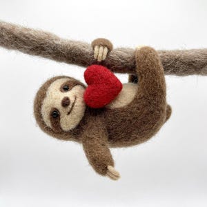 May include: A handcrafted felt sloth holding a red heart, suspended from a brown branch. The sloth features brown and cream-coloured fur and a smiling face. A charming, whimsical decoration.