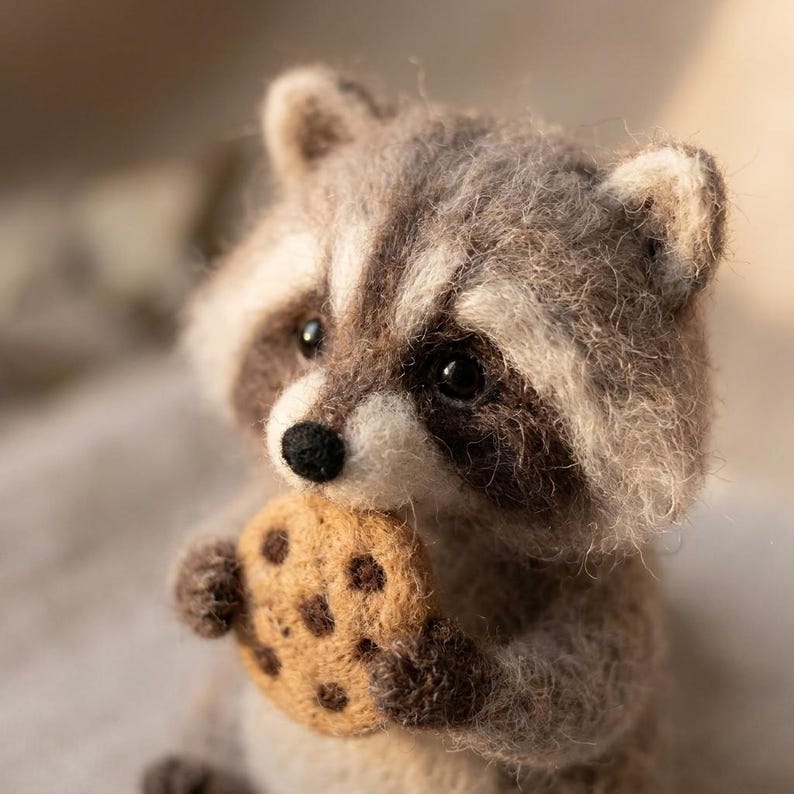 May include: A needle-felted raccoon figurine crafted from fuzzy gray, white, and dark brown wool fibers. The raccoon has black bead eyes, a small black nose, and is holding a round, tan-colored cookie with dark brown chocolate chip spots.
