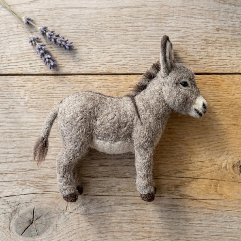 May include: A needle-felted donkey figurine crafted from gray wool with a darker gray mane, tail, and hooves, featuring a light gray underbelly and a small sprig of dried purple lavender flowers positioned near the donkey's head.