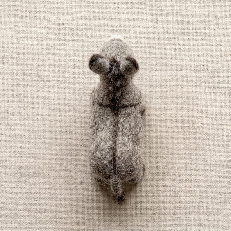 May include: A needle-felted gray donkey figurine viewed from directly above. The figure features a textured, fuzzy wool surface, two upright ears, a dark mane running down the center of the back, and a small tail extending from the rear end.