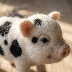 May include: A needle-felted pig figurine crafted from off-white wool fibers with distinct black spots across its body. The small animal sculpture features a soft, fuzzy texture, a light pink snout, and dark, round eyes, capturing a realistic appearance.