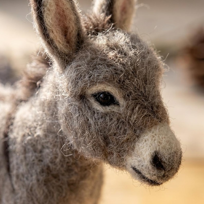 May include: A needle-felted donkey figurine crafted from textured gray and white wool fibers. The donkey features long, upright ears, a soft fuzzy coat, dark eyes, and a distinctively shaped muzzle with a dark nose, capturing a realistic animal form.