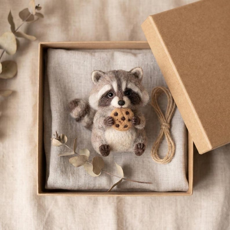 May include: A needle-felted raccoon figurine with grey, white, and black fur, holding a small tan cookie with brown spots. The raccoon is placed inside a square cardboard gift box lined with light grey fabric, accompanied by a piece of twine.